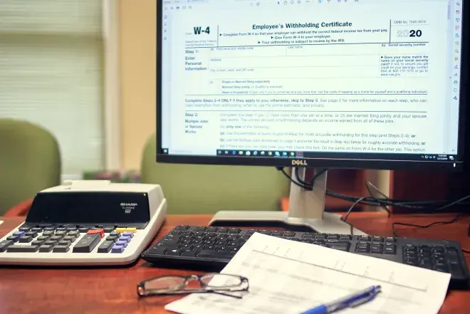 A computer monitor displaying a W-4, with papers, a calculator, and glasses around it.